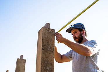 Man wearing baseball cap and sunglasses on building site, using tape measure, Oxfordshire, England