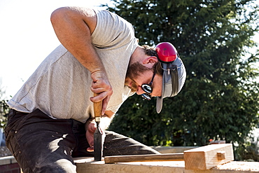 Man wearing baseball cap, sunglasses and ear protectors on building site, working on wooden beam, Oxfordshire, England