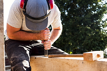 Man wearing baseball cap and ear protectors on building site, working on wooden beam, Oxfordshire, England