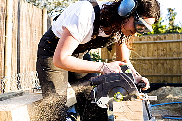 Woman wearing protective goggles and ear protectors holding circular saw, cutting piece of wood on building side, Oxfordshire, England