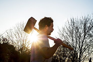 Bearded man walking outdoors at sunset, carrying large wooden mallet on his shoulder, Oxfordshire, England