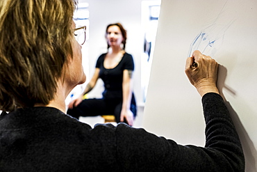 Close up of woman standing at easel, drawing human hand, artist's model sitting in the background. A life drawing class with a model, Oxfordshire, England