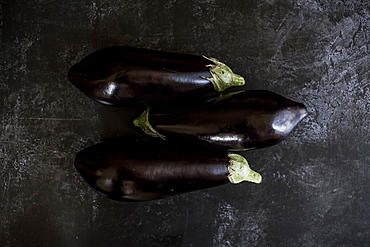 Three glossy purple aubergine egg plant vegetables on a grey slate background, England