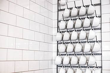 Close up of white cappuccino mugs hanging on a metal rack on white tiled wall