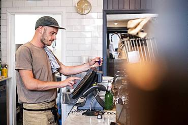 Bearded man wearing baseball cap standing at a till in a restaurant