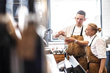 Male and female chef wearing brown aprons standing at a counter, checking an order