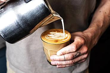 Close up of person with tattooed finger pouring milk from metal jug into glass of cafe latte