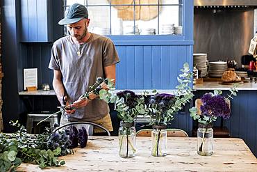 Man wearing baseball cap standing at a table, cutting thistles for flower arrangements in large glass jars