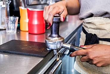 Woman barista preparing coffee beans grounds, tamping them down into a grounds holder in a cafe