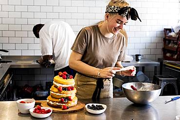 A cook working in a commercial kitchen assembling a layered sponge cake with fresh fruit