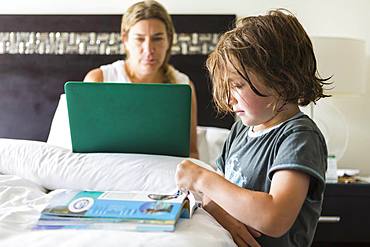 5 year old boy looking at book in hotel room as mom works on laptop, Grand Cayman, Cayman Islands