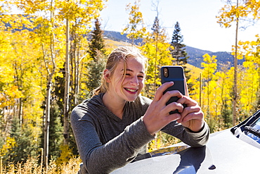 13 year old girl taking pictures with her smart phone, looking at autumn aspen trees