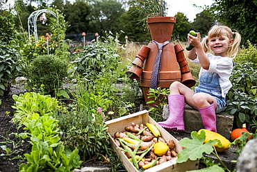 Smiling blond girl sitting in garden next to terracotta scarecrow, picking fresh vegetables