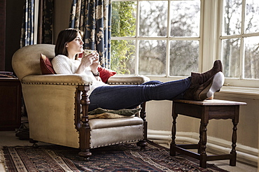 A young woman relaxing at home, with her feet up, having a cup of tea, Ringwood, Hampshire, England