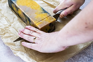 High angle close up person cutting yellow and black homemade bar of soap with kitchen knife