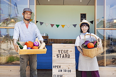 Japanese man and woman standing outside a farm shop, holding crate and basket with fresh vegetables, looking at camera, Kyushu, Japan