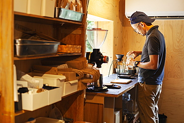 Japanese man wearing baseball cap and glasses standing in an Eco Cafe, preparing cup of coffee, Kyushu, Japan