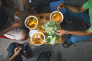 High angle close up of three men tucking into bowls of noodles for breakfast in a small alleyway, Vietnam