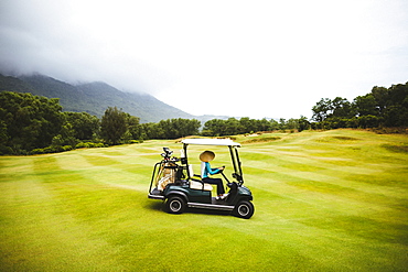 Woman wearing traditional straw hat driving golf cart on the green of a golf course under a cloudy sky