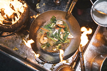 High angle close up of stir fry with prawns and green vegetables in stainless steel wok over flame of charcoal grill, Thailand