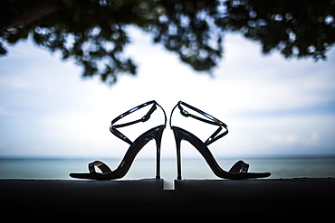 Close up of pair of shiny stiletto sandals, tree foliage and ocean in background, Thailand