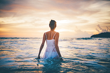 Rear view of young woman wearing a white dress walking in the ocean at sunset, Thailand