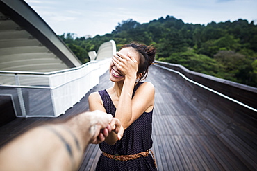 Smiling young woman standing on a bridge, covering her face, holding man's hand, Singapore