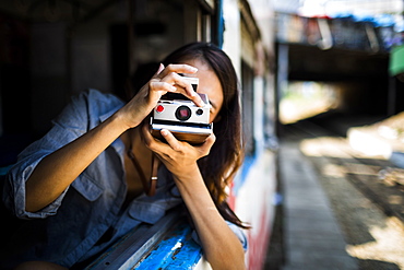 Young woman riding on a train, looking out of window, taking picture with old Polaroid camera, Myanmar