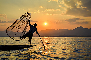 A fisherman balancing on one leg on a boat, holding a large fishing basket, fishing in the traditional way on Lake Inle at sunset, Myanmar