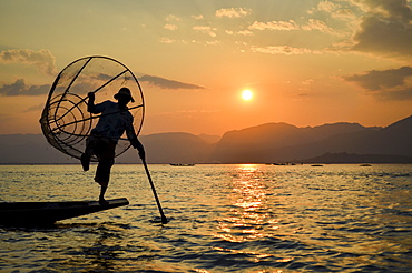 Traditional fisherman balancing on one leg on a boat, holding fishing basket, fishing on Lake Inle at sunset, Myanmar