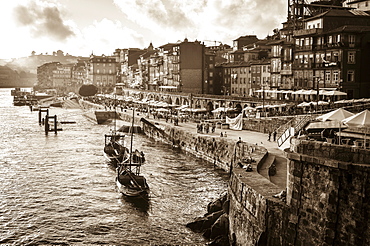 Port boats and freight barges moored beside a waterfront wall, and people on the promenade. Historic buildings, Porto, Portugal