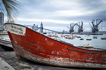 An old boat with a red shabby hull beached on the shore by the water in Lisbon. Shipping cranes and a large modern road bridge in the distance, Lisbon, Portugal