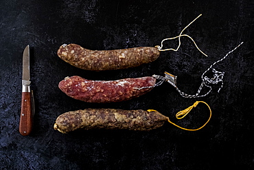 High angle close up of knife and selection of three salamis on black background, United Kingdom