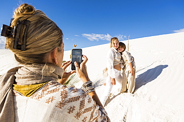 A woman taking picture of her children with a smart phone in white sand dunes landscape under blue sky, White Sands National Monument, New Mexico, United States