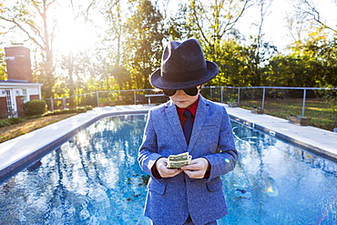 Boy in a suit and dark glasses standing on the edge of water counting dollar bills.