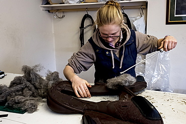 Female saddler standing in workshop, stuffing leather saddle with horse hair, Berkshire, United Kingdom