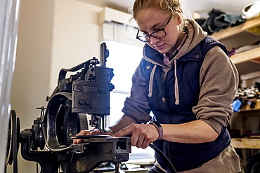 Female saddler standing in workshop, sewing leather strap on saddlery sewing machine, Berkshire, United Kingdom