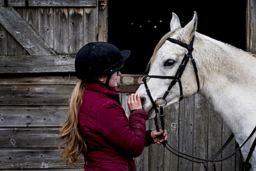 Young woman standing outside stable, holding white Cob horse by it's reins, Berkshire, United Kingdom