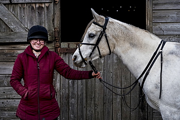 Young woman standing outside stable, holding white Cob horse by it's reins, Berkshire, United Kingdom