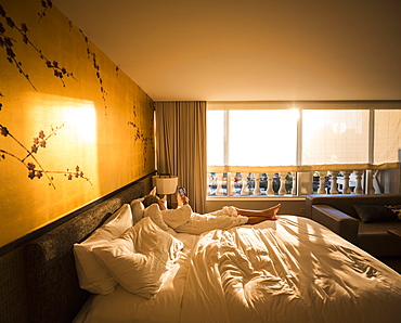 Adult woman executive lying on hotel bed in early morning sunlight, looking at smart phone, United States