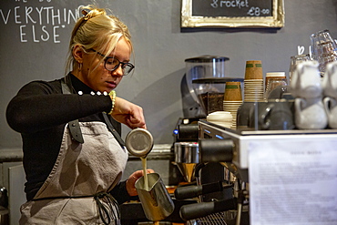 Blond woman wearing glasses and apron standing at espresso machine in a cafe, pouring milk into metal jug