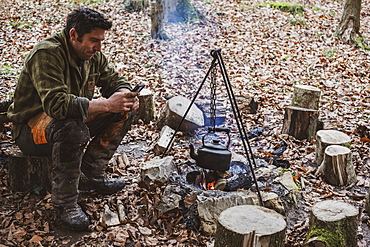 Man sitting by a camp fire in a forest, boiling kettle of water, Devon, United Kingdom