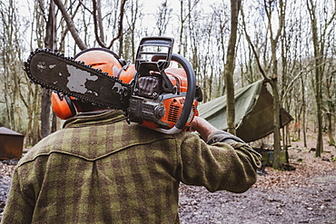 Rear view of man wearing safety gear walking through forest carrying chainsaw on his shoulder, Devon, United Kingdom