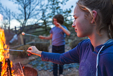 A teenage girl making smores with her brother over a fire in a garden at dusk