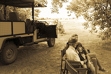 Three generations of a family seated in folding chairs by a safari jeep, under trees, monochrome, Moremi Game Reserve, Botswana