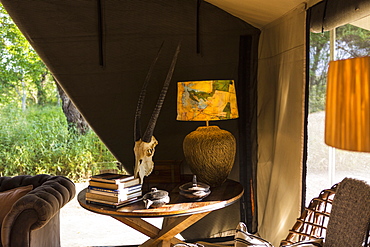 The interior of a tent in a safari camp with electric lamp and table, chairs and sofa, tent sides rolled up