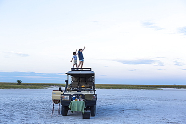 A brother and sister standing on top of a safari vehicle at dusk in a salt pan landscape, Makgadikgadi Pan National Park, Botswana