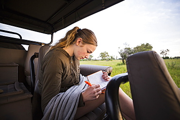 A teenage girl writing in her journal in a safari vehicle in the bush, Botswana