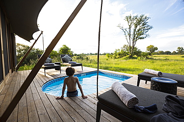 A boy in a swimming pool at a safari camp, looking out over the landscape, Botswana