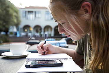 A teenage girl using pen and paper making notes, looking at a smart phone, writing a diary or doing homework